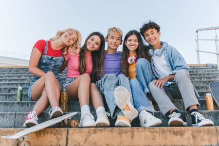 Téléchargez les photos : Portrait d'adolescents multiraciaux souriants assis dans des escaliers regardant une caméra dans une rue urbaine. Amis heureux embrasser ensemble en plein air Amitié, concept de diversité - en image libre de droit