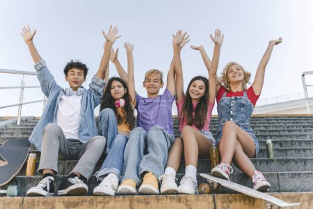 Téléchargez les photos : Groupe d'amis heureux, les adolescents garçons et les filles en t-shirts colorés tenant la main vers le haut célébration vacances d'été assis sur les escaliers - en image libre de droit