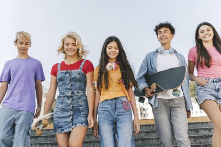 Foto de Grupo de amigos adolescentes multirraciales sonrientes que usan camisetas coloridas hablando, comunicándose, sosteniendo patinetas caminando por la calle. Feliz elegante chicos y chicas al aire libre. Concepto de amistad - Imagen libre de derechos