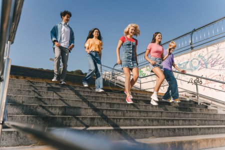 Foto de Grupo de amigos adolescentes multirraciales sonrientes vistiendo colorida camiseta hablando, comunicación caminando por la calle. Feliz elegante chicos y chicas al aire libre. Concepto de amistad - Imagen libre de derechos
