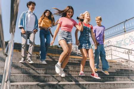 Photo for Group of smiling friends, multiracial teenagers wearing colorful casual clothes running on the street, having fun. Happy stylish boys and girls outdoors. Friendship, positive lifestyle, summer - Royalty Free Image
