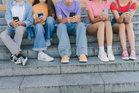 Photo for Cropped picture of group teenagers holding mobile phones watching video, communication online, chatting sitting on the street. Technology, social media concept - Royalty Free Image