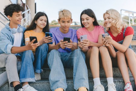 Téléchargez les photos : Groupe d'amis souriants, adolescents multiraciaux tenant des téléphones mobiles regardant la vidéo, la communication en ligne, jouant au jeu mobile, parlant assis sur les escaliers. Technologie, concept de médias sociaux - en image libre de droit