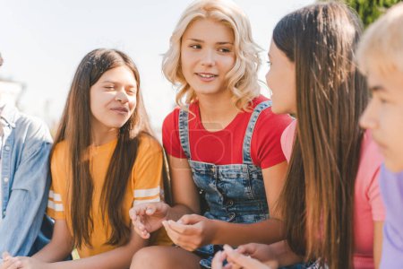 Foto de Retrato de adolescentes sonrientes, chicas con elegantes camisetas de colores hablando, sentadas juntas. Amigos positivos de la escuela reunidos en la calle, al aire libre. Concepto de amistad - Imagen libre de derechos