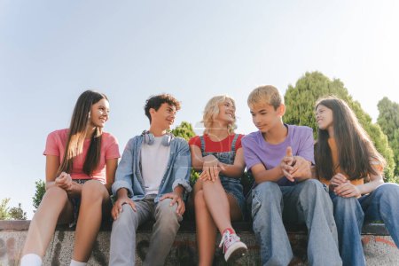 Téléchargez les photos : Groupe d'adolescents souriants parlant, communication ensemble assis dans le parc. Concept d'été - en image libre de droit