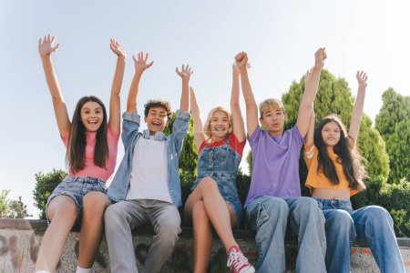 Téléchargez les photos : Groupe d'amis émotionnels heureux, adolescents multiraciaux garçons et filles en t-shirts colorés tenant la main vers le haut célébration vacances d'été assis dans la rue, en s'amusant. Style de vie positif, été - en image libre de droit