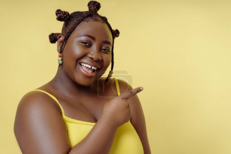 Foto de Retrato de una mujer africana sonriente mirando a la cámara aislada sobre fondo amarillo, señalando con el dedo el espacio de copia - Imagen libre de derechos