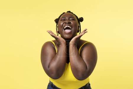 Téléchargez les photos : Portrait de femme nigériane souriante positive avec une coiffure élégante et les yeux fermés touchant visage émotionnel isolé sur fond jaune. Attrayant modèle à la mode du corps féminin positif, beauté naturelle. Concept de modèle Happy plus size - en image libre de droit