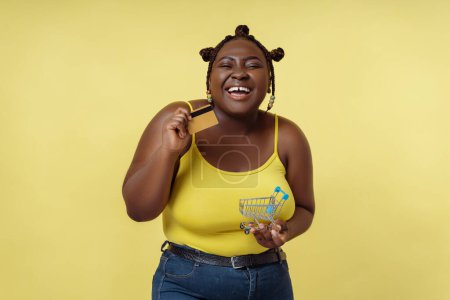Photo for Portrait of smiling African woman holding credit card, looking at camera isolated on yellow background. Smiling female with stylish hairstyle, online banking - Royalty Free Image