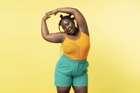 Téléchargez les photos : Souriant femme africaine attrayante faire des exercices en regardant la caméra isolée sur fond jaune. Concept taille plus, mode de vie sain - en image libre de droit
