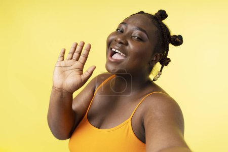 Foto de Auténtica mujer africana más tamaño positivo tomando selfie grabación video saludando mano aislada sobre fondo amarillo. Atractiva influenciadora nigeriana posando para la foto - Imagen libre de derechos