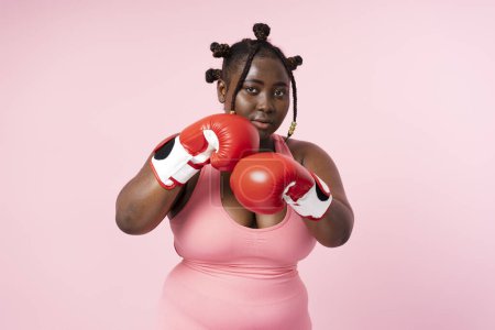 Téléchargez les photos : Portrait de femme africaine portant des gants de boxe rouges, isolée sur fond rose. Entraînement féminin sérieux de plus de taille, motivation, concept sain de mode de vie - en image libre de droit