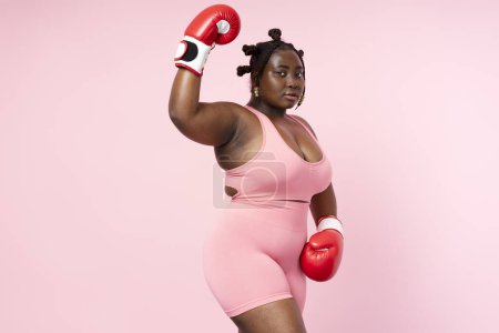 Photo for Pensive beautiful Nigerian woman wearing sports uniform red boxing gloves looking at camera isolated on pink background. Authentic body positive African model posing for picture, copy space - Royalty Free Image