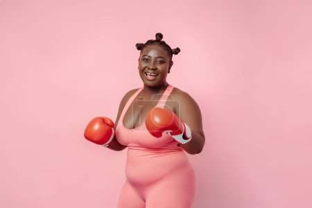Foto de Retrato de una mujer africana con cuerpo positivo y guantes de boxeo rojos aislados sobre fondo rosa. Entrenamiento femenino, motivación, hobby - Imagen libre de derechos
