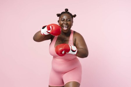 Téléchargez les photos : Souriante femme afro-américaine positive portant un uniforme de sport, des gants de boxe rouges regardant la caméra isolée sur fond rose. Formation de sportives nigérianes, soins de santé, mode de vie - en image libre de droit