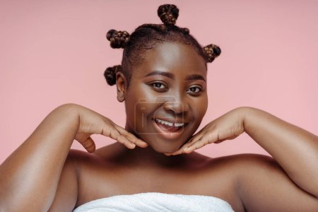 Foto de Sonriente mujer africana alegre en toalla después de la ducha mirando en espejo tocando la cara aislada sobre fondo rosa. Concepto de cuidado de la piel, estilo de vida saludable - Imagen libre de derechos