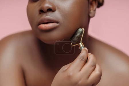 Photo for Closeup of beautiful african american woman doing face roller massage, looking in mirror isolated on pink background. Natural beauty, skin care, body positivity concept - Royalty Free Image