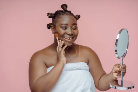 Téléchargez les photos : Portrait de belle femme afro-américaine avec coiffure élégante après la douche dans une serviette, tenant miroir, regardant miroir isolé sur fond rose. Soins de la peau, procédures beauté - en image libre de droit
