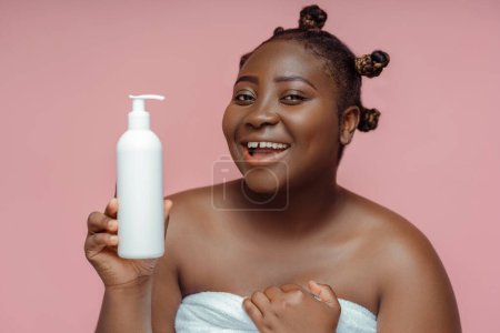 Téléchargez les photos : Sourire femme africaine positive dans la serviette après la douche tenant hydratant isolé sur fond rose. Concept de soins de la peau et du corps - en image libre de droit