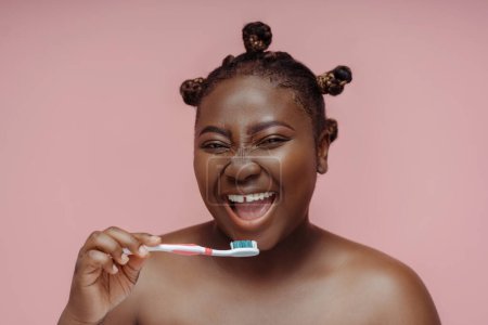 Téléchargez les photos : Belle femme africaine regardant dans le miroir, brossant les dents avec une brosse à dents isolée sur fond rose. Concept de soins dentaires et de santé - en image libre de droit