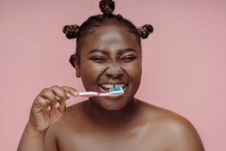 Foto de Mujer afroamericana feliz mirándose en el espejo, cepillándose los dientes con cepillo de dientes aislado sobre fondo rosa. concepto de estilo de vida saludable - Imagen libre de derechos