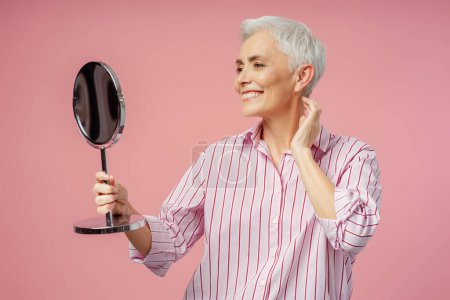 Téléchargez les photos : Belle femme âgée souriante aux cheveux gris, maquillage élégant regardant dans un miroir isolé sur fond rose. Beauté naturelle, routine matinale, mode de vie sain concept - en image libre de droit