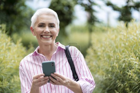 Téléchargez les photos : Femme mûre souriante tenant un téléphone mobile, faisant du shopping en ligne dans la rue, copiant de l'espace. Belle femme âgée aux cheveux gris vérifiant le courrier. Concept technologique - en image libre de droit