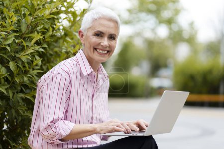 Portrait of successful senior businesswoman using laptop, sitting on bench outdoors. Attractive mature freelancer working online, workplace. Business concept