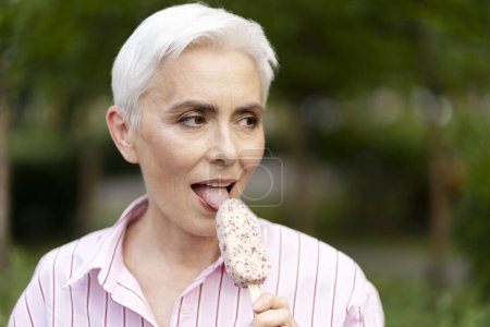 Photo for Beautiful elderly woman eating ice cream on street looking away. Female enjoying dessert outdoors, tasty concept - Royalty Free Image