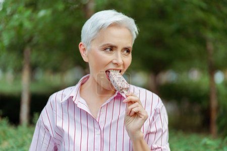 Téléchargez les photos : Heureuse femme aux cheveux gris élégant mangeant une délicieuse crème glacée et regardant la distance debout dans le parc. Vacances d'été, concept de style de vie positif - en image libre de droit
