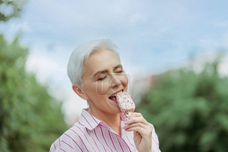 Photo for Portrait of happy stylish gray haired woman eating tasty ice cream with close eyes standing in park. Summer vacation, positive lifestyle concept - Royalty Free Image