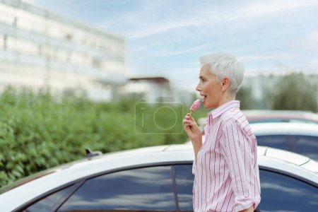 Foto de Feliz mujer de pelo gris elegante comiendo sabroso helado mientras disfruta de caminar en el parque. Estilo de vida positivo y sabroso concepto de comida - Imagen libre de derechos