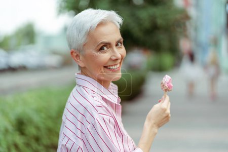 Photo for Cheerful stylish gray haired woman looking at camera while eating tasty ice cream in park. Positive lifestyle and tasty food concept - Royalty Free Image