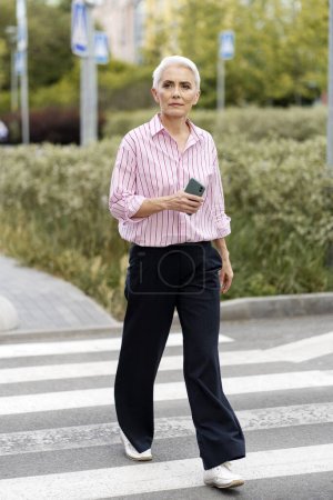Photo for Serious senior woman in casual clothes holding mobile phone walking down street. Attractive businesswoman using smartphone. Technology concept - Royalty Free Image