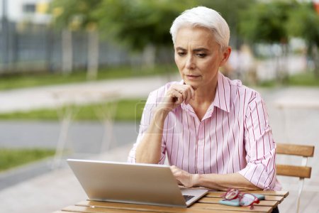 Photo for Portrait of serious senior woman using laptop, sitting at table on street, copy space. Successful businesswoman wearing pink shirt working remotely. ?oncept of modern business - Royalty Free Image