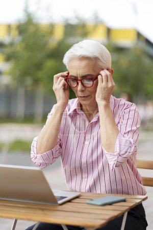 Foto de Retrato de una mujer mayor con anteojos rojos usando laptop, sentada al aire libre. Anciano freelancer trabajando en línea, trabajo remoto. Concepto de negocio exitoso - Imagen libre de derechos