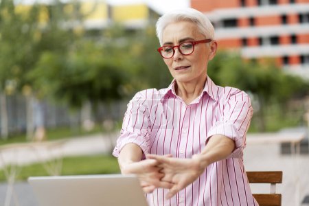 Foto de Hermosa mujer jubilada con elegantes gafas rojas y camisa casual, utilizando el ordenador portátil, estirando las manos. Empresaria exitosa, descanso, concepto de trabajo en línea exitoso - Imagen libre de derechos
