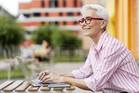 Foto de Retrato de una mujer mayor positiva sonriente que usa gafas rojas modernas, trabaja en la computadora, usa una computadora portátil al aire libre, mira hacia otro lado, copia el espacio. Autónomo exitoso, lugar de trabajo. Concepto de trabajo remoto - Imagen libre de derechos
