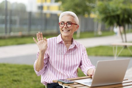 Téléchargez les photos : Femme âgée positive souriante portant des lunettes rouges élégantes et des vêtements décontractés à l'aide d'un ordinateur portable à l'extérieur, agitant la main, regardant ailleurs. ? l'acceptation du travail indépendant, du travail à distance - en image libre de droit