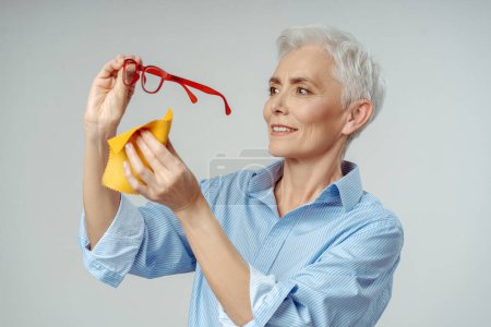 Foto de Mujer mayor sonriente confiada limpiando nuevas gafas rojas con estilo aisladas sobre fondo gris, concepto de visión. Atractivo feliz gris mujer de negocios de pelo limpieza de ropa de los ojos - Imagen libre de derechos