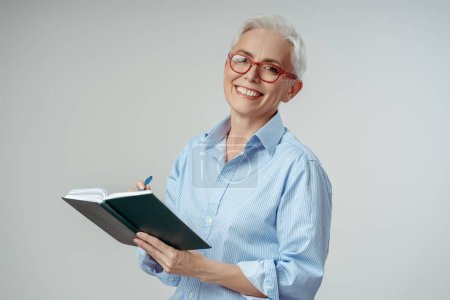 Foto de Atractiva mujer de pelo gris con gafas con estilo libro de lectura, proyecto de planificación aislado sobre fondo gris. Estudiante de último año estudiando, aprendiendo el idioma, copiando espacio. Concepto educativo - Imagen libre de derechos