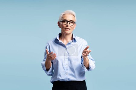 Photo for Serious gray haired female teacher wearing eyeglasses and stylish blue shirt explaining, looking at camera. Attractive businesswoman taking part in video conference. Concept of successful education - Royalty Free Image