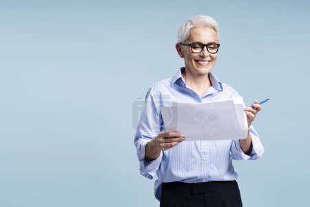 Photo for Confident smiling mature businessman wearing glasses holding business graph isolated on blue background, copy space. Beautiful teacher working with documents. Successful education concept - Royalty Free Image