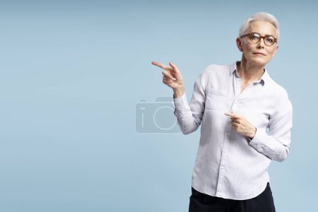Foto de Atractiva mujer mayor con gafas y ropa casual señalando los dedos en el espacio de copia aislado en el fondo azul. Mujer de negocios de pelo gris apuntando al espacio vacío, publicidad - Imagen libre de derechos