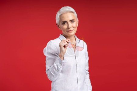 Photo for Portrait of attractive gray haired woman holding sunglasses looking at camera isolated on red background. Beautiful business woman in stylish pink shirt, successful employee. Concept of business - Royalty Free Image