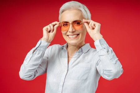Foto de Retrato de una mujer mayor sonriente probándose unas gafas de sol con estilo naranja mirando a la cámara sobre el fondo rojo. Concepto de personas exitosas - Imagen libre de derechos