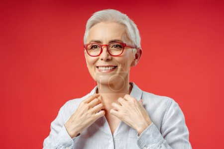 Foto de Mujer mayor de pelo gris sonriente que usa ropa de ojo elegante aislada sobre fondo rojo. Atractiva mujer de negocios, CEO, gerente mirando hacia otro lado. Negocios exitosos - Imagen libre de derechos