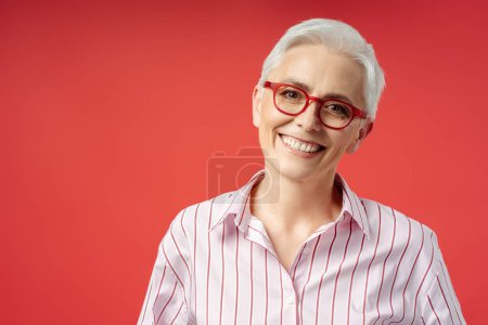 Foto de Retrato de mujer mayor feliz con gafas rojas elegantes aisladas sobre fondo rojo, concepto de visión. Atractiva sonriente mujer de negocios de pelo gris, gerente seguro mirando a la cámara - Imagen libre de derechos