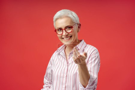 Foto de Retrato de mujer mayor de pelo gris sonriente con gafas elegantes coqueteando cámara aislada sobre fondo rojo, concepto de visión. Atractiva empresaria posando - Imagen libre de derechos