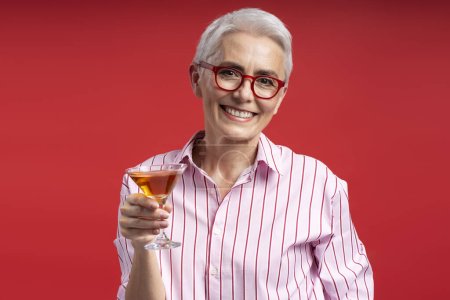 Foto de Atractiva mujer mayor de pelo gris con elegantes gafas rojas sosteniendo vidrio con bebida aislada sobre fondo rojo. Sonriente anciana mirando a la cámara - Imagen libre de derechos